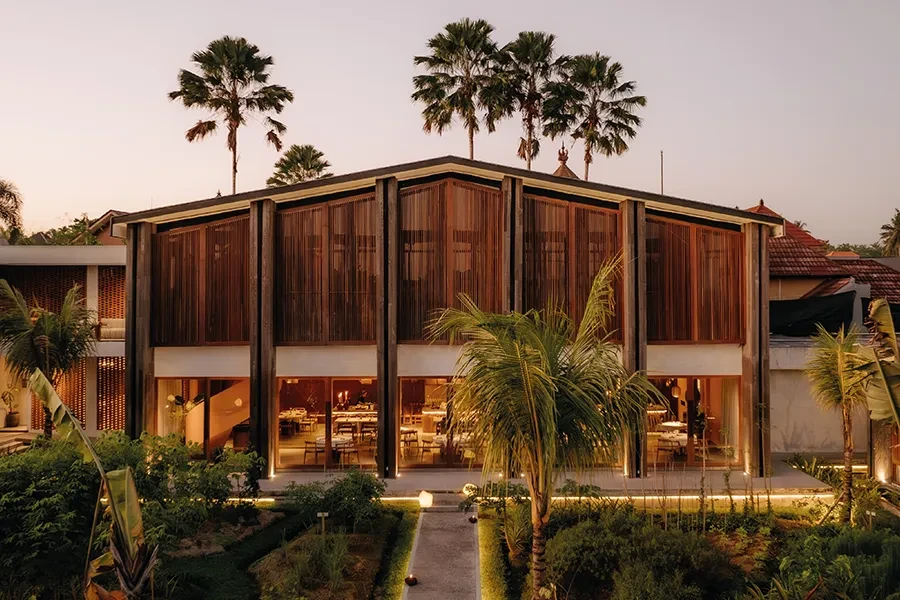 The illuminated wood and glass facade of Syrco BASE restaurant at dusk, showcasing the warm interior dining area and lush garden landscaping.